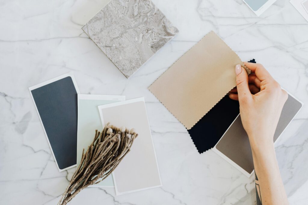 Hands holding fabric swatches with color samples on a marble surface for interior design inspiration.