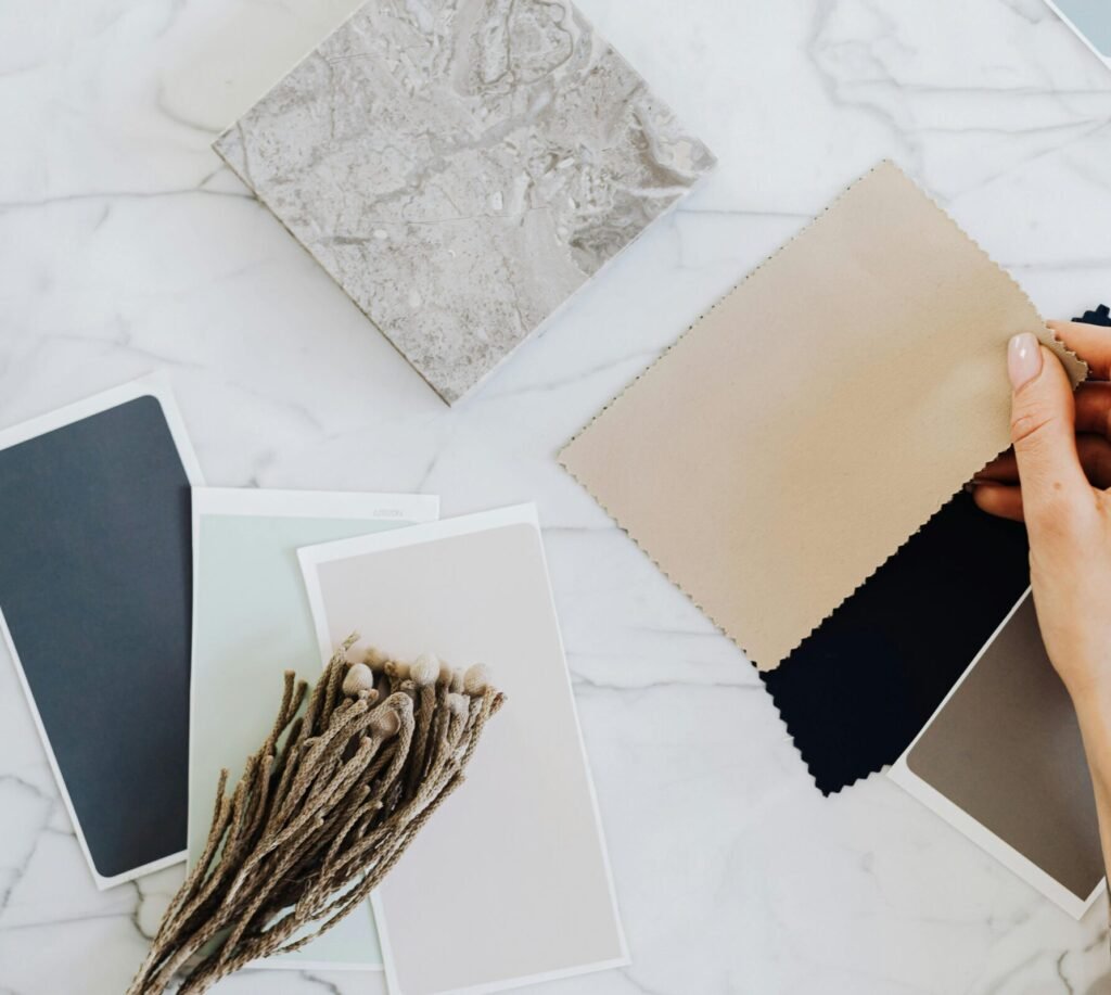 Hands holding fabric swatches with color samples on a marble surface for interior design inspiration.