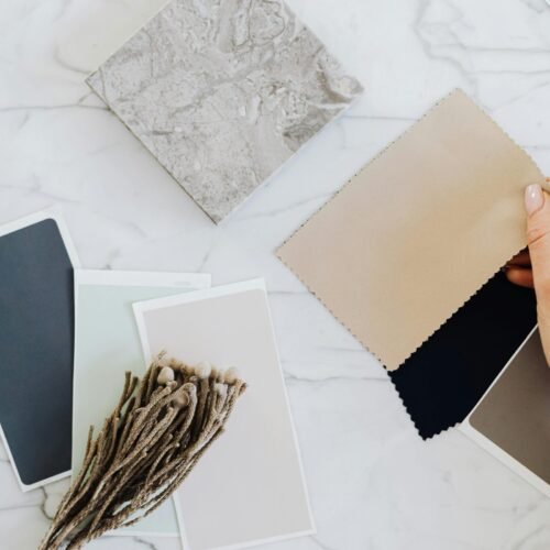 Hands holding fabric swatches with color samples on a marble surface for interior design inspiration.